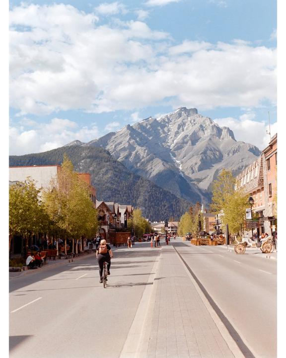 A person rides a bicycle down a street lined with buildings and trees, with a large mountain in the background under a cloudy sky.