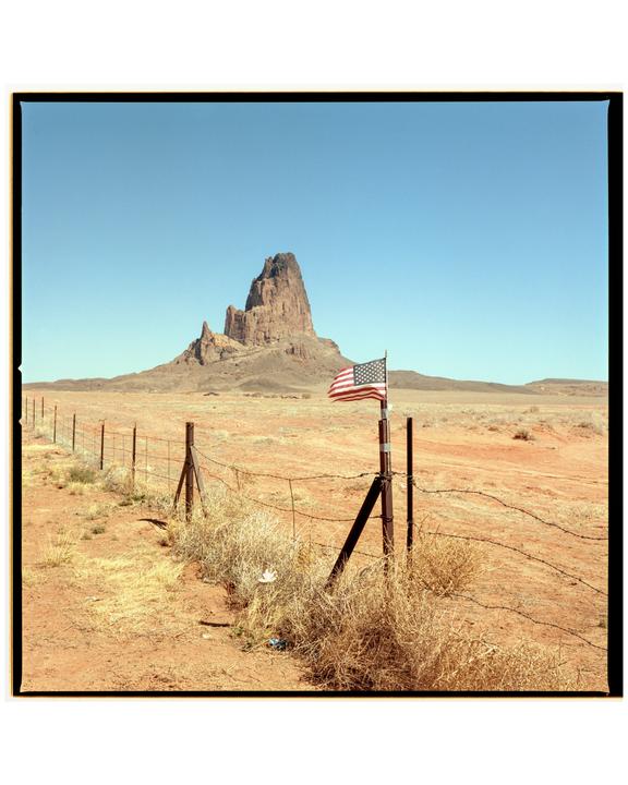 An American flag is attached to a fence post in a desert landscape with a large rock formation in the background.