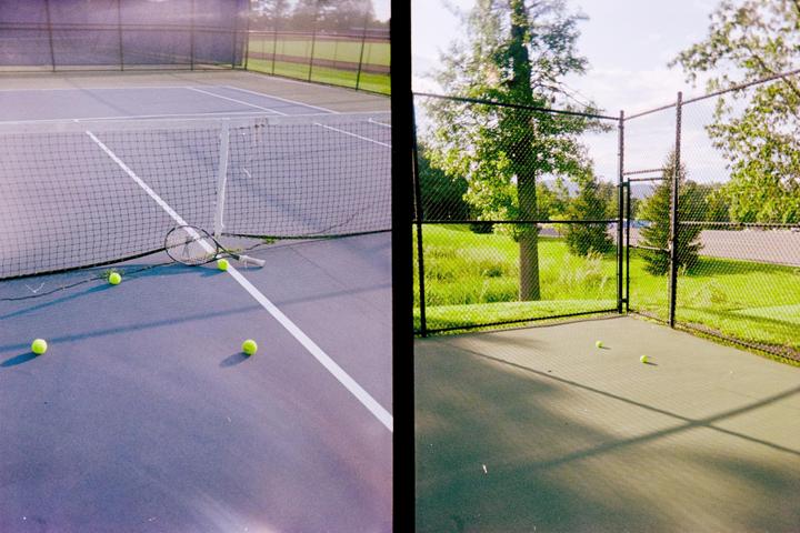 A tennis racket and several tennis balls are scattered across a tennis court next to a net and a fence.