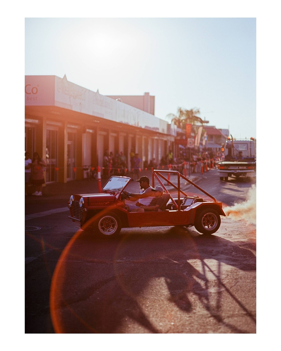 A man drives a red vehicle with a child in the passenger seat, leaving a trail of orange smoke behind it on a street.