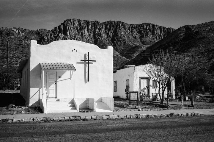 A white adobe church with a cross on its facade stands next to another building, with mountains in the background.