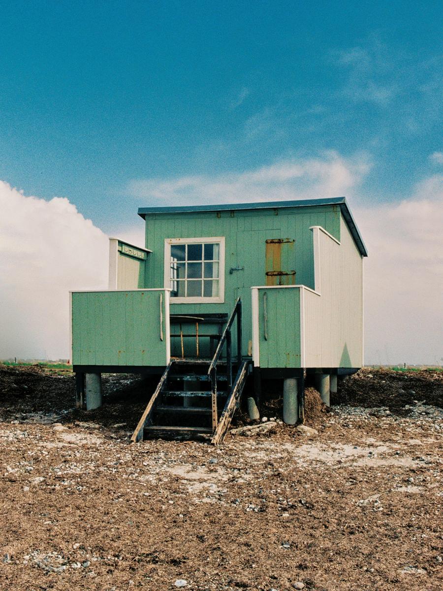 A small green wooden house with a white window and a dark staircase stands on a rocky, brown ground under a blue sky with white clouds.