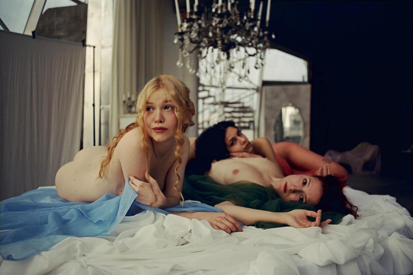 Three women are reclining on a bed with white sheets and colorful fabric.