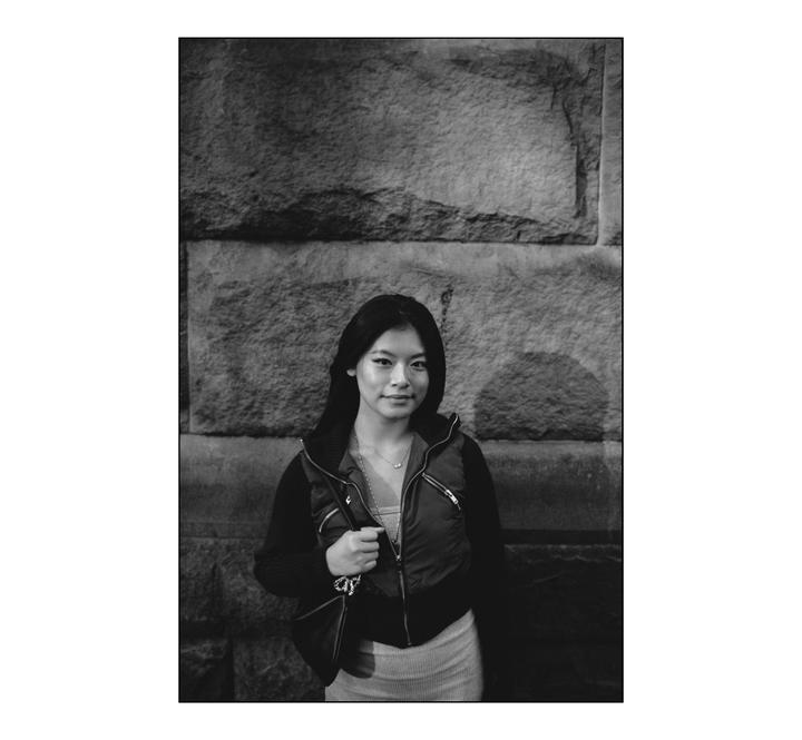 A woman stands in front of a stone wall, looking at the viewer.