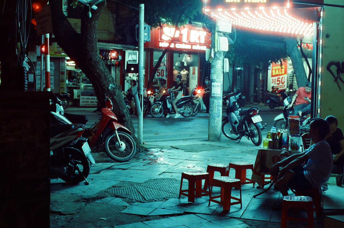 A street scene at night shows motorbikes parked along a sidewalk with people sitting at a small table with red stools under bright lights.