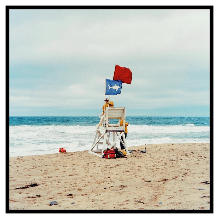 Two people stand on and next to a lifeguard chair on a sandy beach with two flags flying above it.