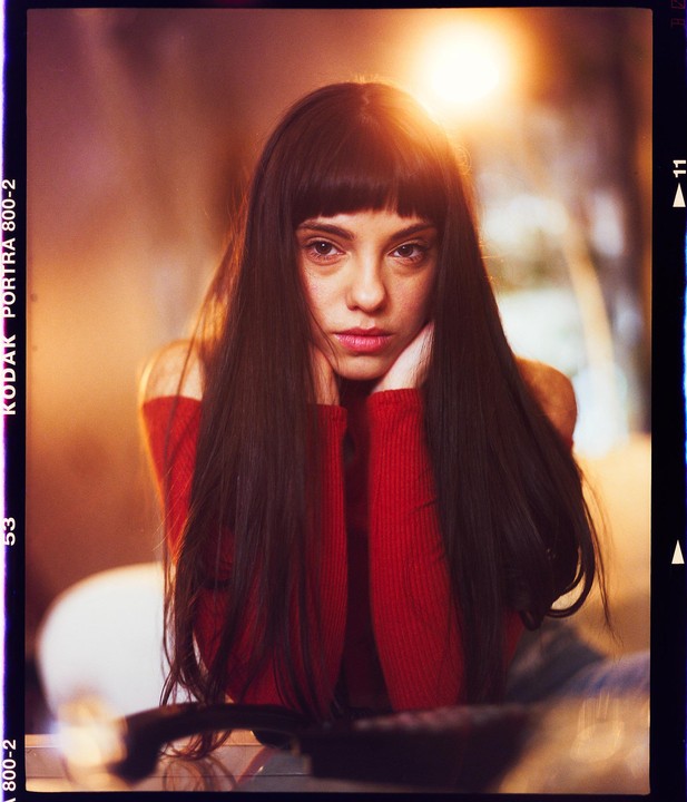 A woman with long dark hair and bangs wears a red sweater and rests her chin on her hands.