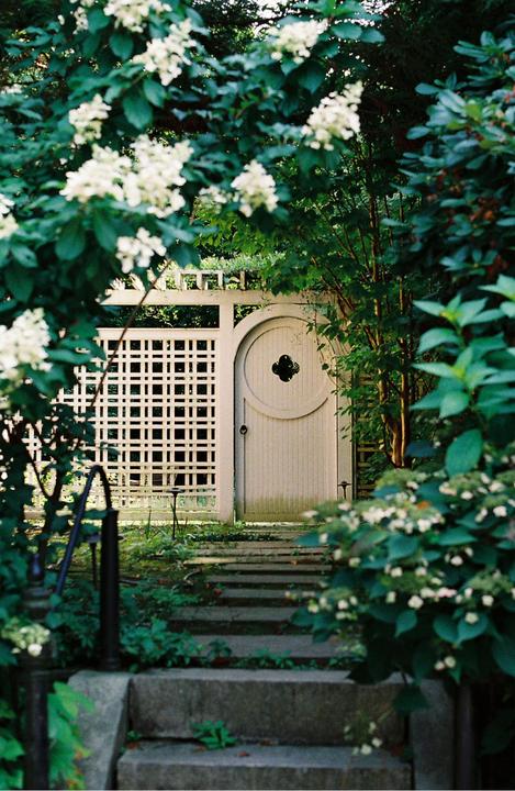 A white garden gate with a circular design is framed by green foliage and white flowers, with stone steps leading up to it.