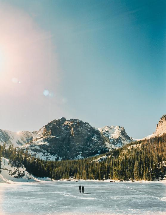 Two people walk across a frozen lake surrounded by snow-covered mountains and a forest.