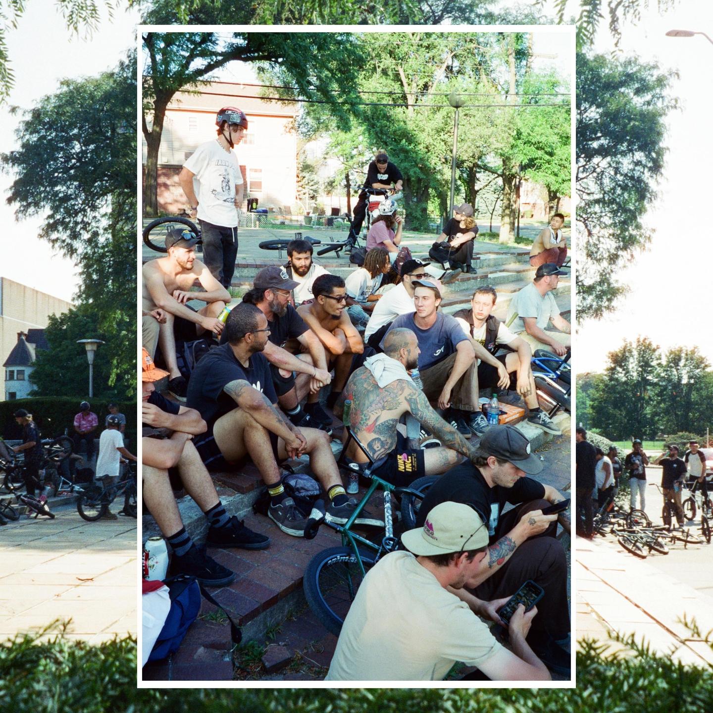 A group of men are gathered on steps and on the ground with bicycles in a park.