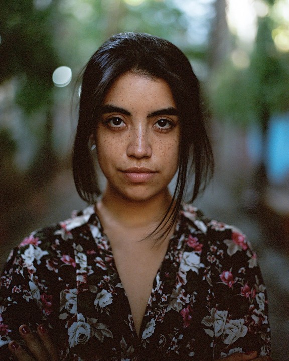 A woman with dark hair and freckles looks directly at the viewer.