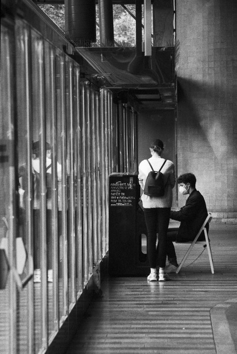 A woman stands next to a man sitting on a folding chair, both facing away from the camera, with a reflective barrier to their left.