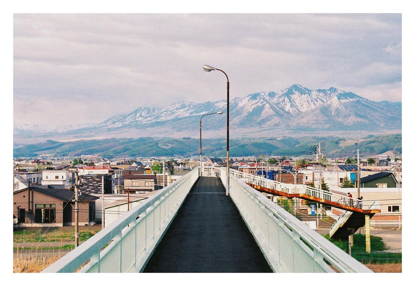 A long pedestrian bridge leads to a town with mountains in the background.