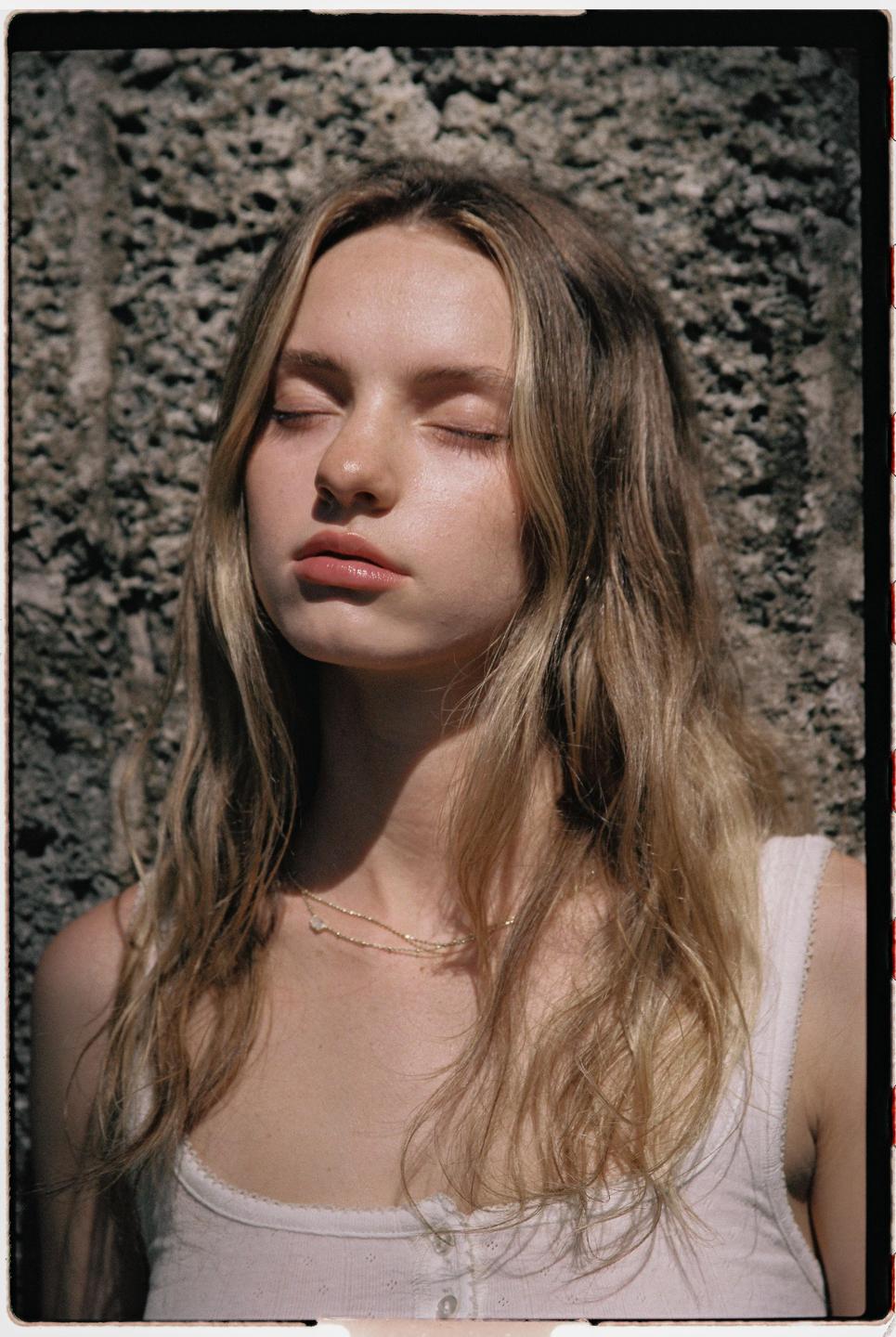 A young woman with closed eyes and long blonde hair stands in front of a textured wall.