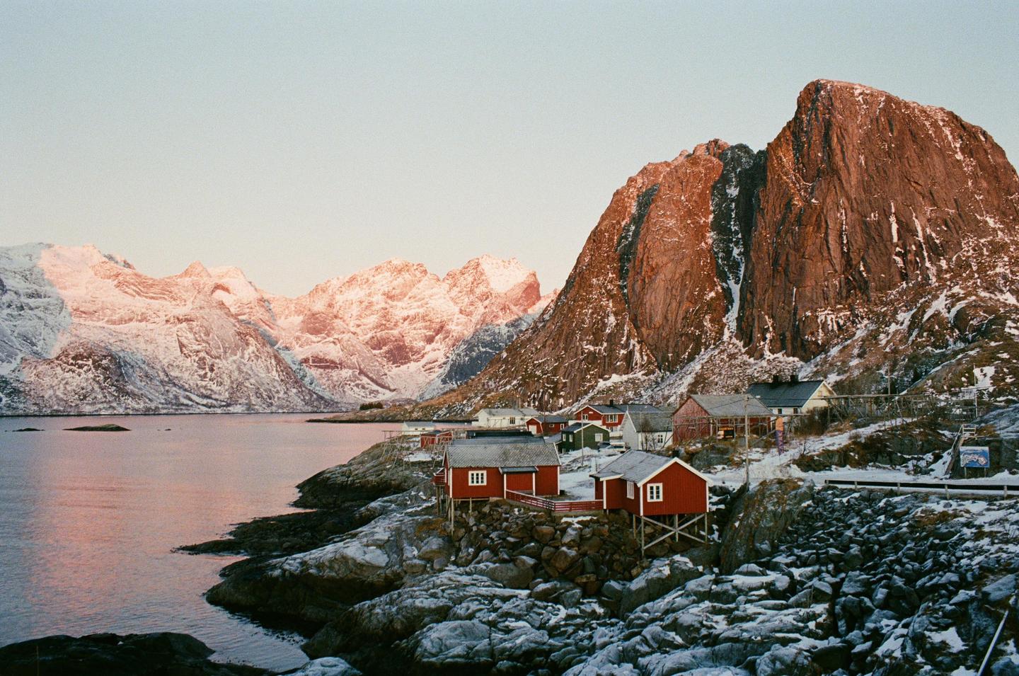A small village with red and white houses sits on a rocky, snow-covered shore next to a body of water, with large, snow-capped mountains in the background.