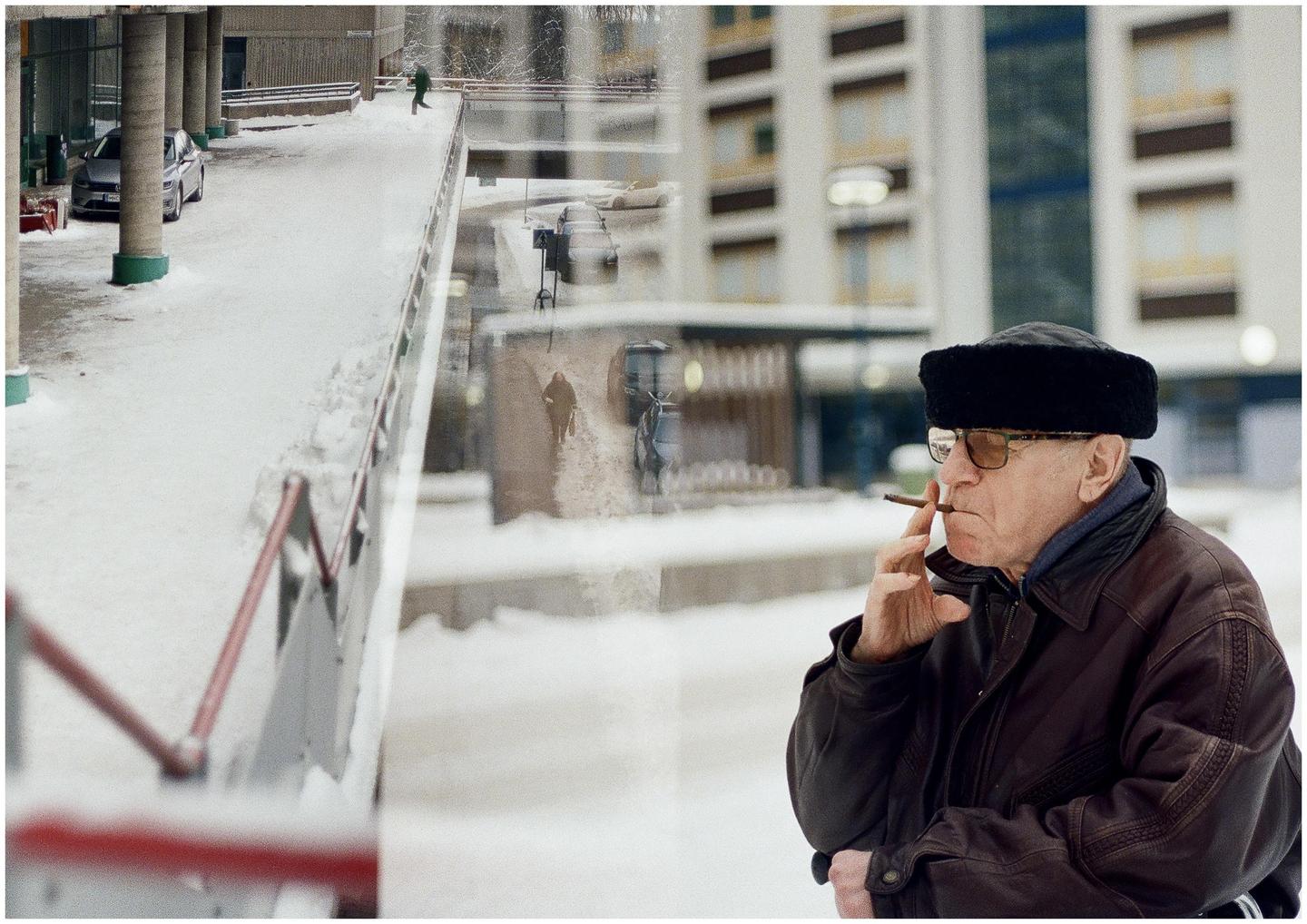 A man smoking a cigar is double exposed over a snowy urban landscape with a car and people.