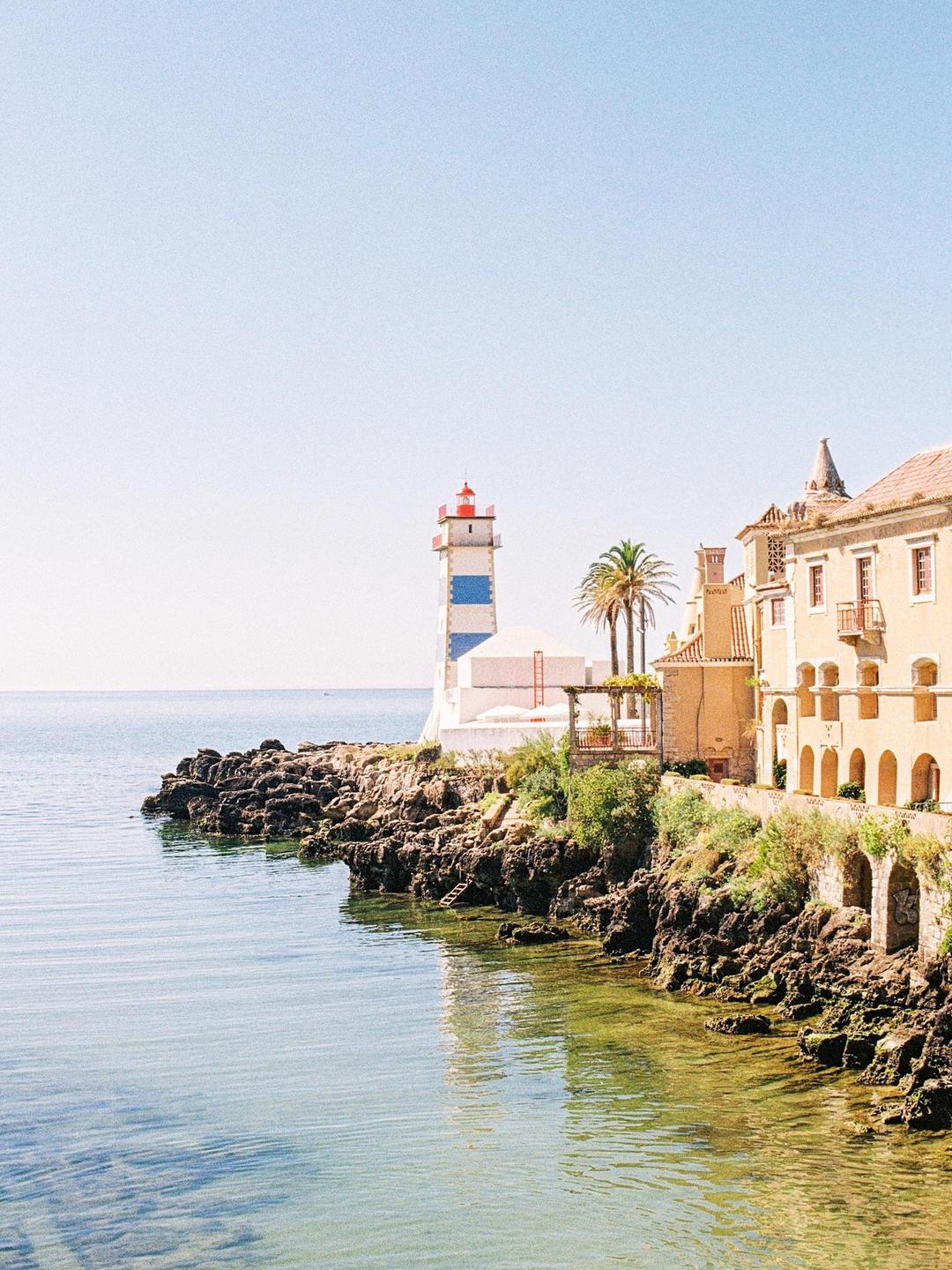 A lighthouse stands on a rocky coast next to buildings and a palm tree.