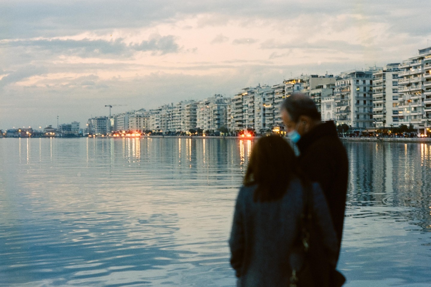 A man and a woman stand by a body of water with a city skyline in the background at dusk.