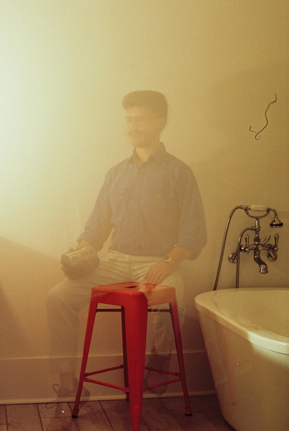 A man sits on a red stool next to a bathtub, holding a can.
