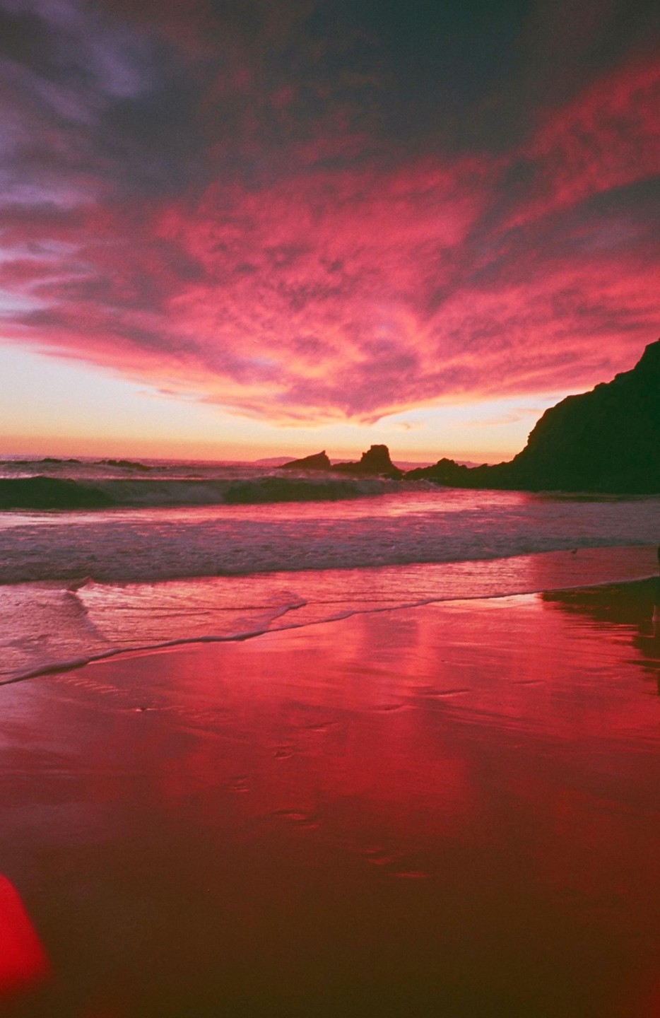 A vibrant pink and orange sunset illuminates the sky and reflects on the wet sand of a beach with waves crashing near dark cliffs.