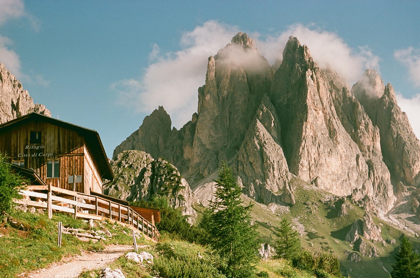 A wooden mountain hut with a fence sits on a grassy slope with towering, rocky mountains in the background, partially obscured by clouds.