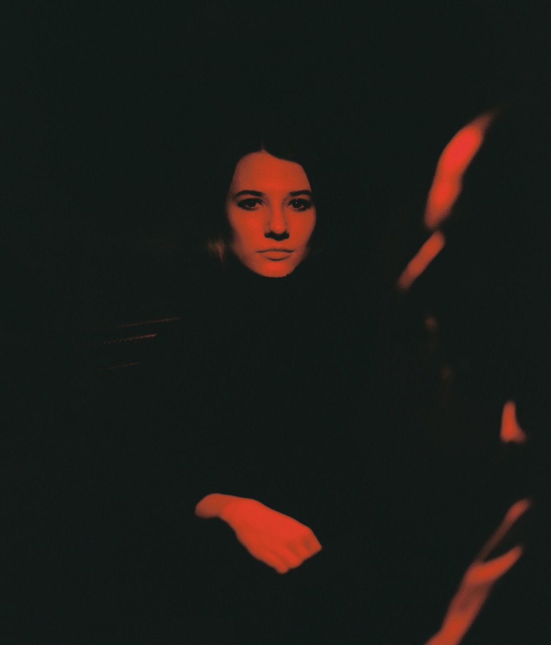 A woman's face and hand are illuminated by red light in a dark setting, with a blurred reflection visible on the right.