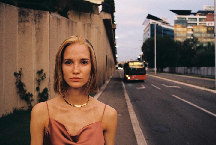 A woman with blonde hair and a pink top stands on a sidewalk next to a road with a bus and buildings in the background.