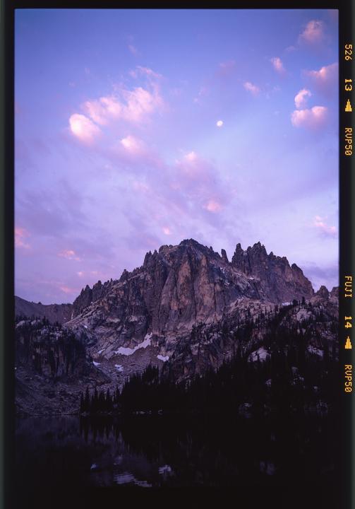 A mountain range with snow and trees is reflected in a lake under a purple and pink sky with a moon and clouds.