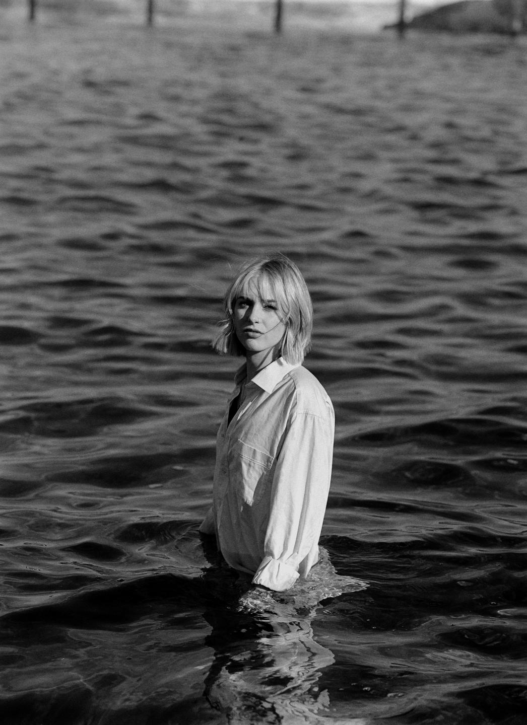A woman in a white shirt stands in the water, looking towards the viewer.