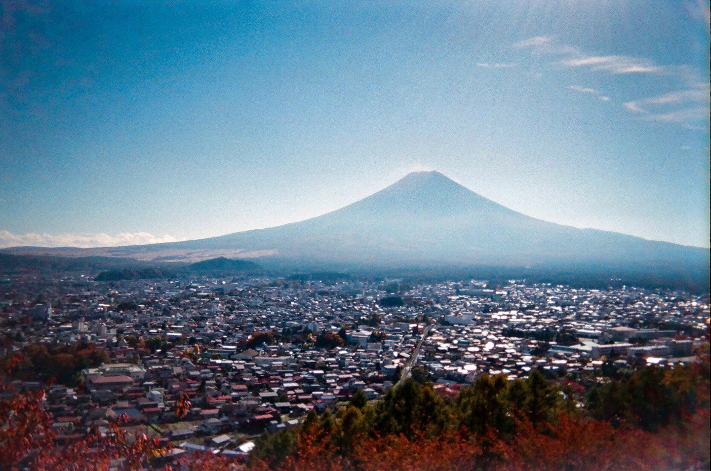 Mount Fuji rises above a city with red foliage in the foreground.