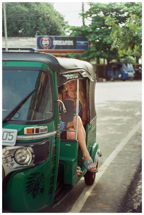 A woman with long blonde hair sits in the passenger seat of a green tuk-tuk on a street.