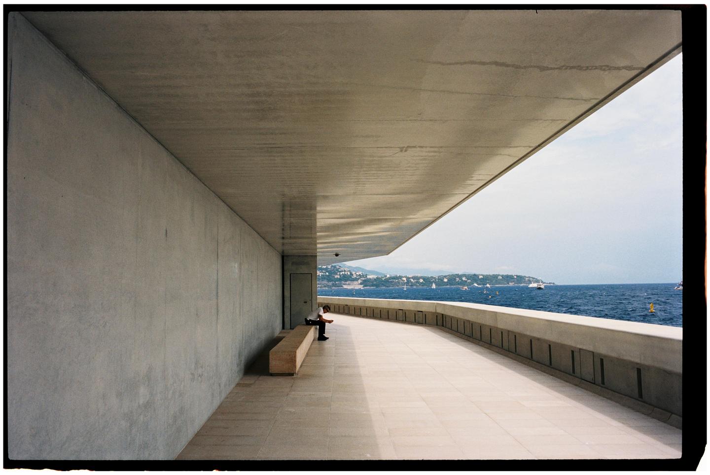 A man sits alone on a bench on a concrete walkway overlooking the sea with boats and a distant coastline.