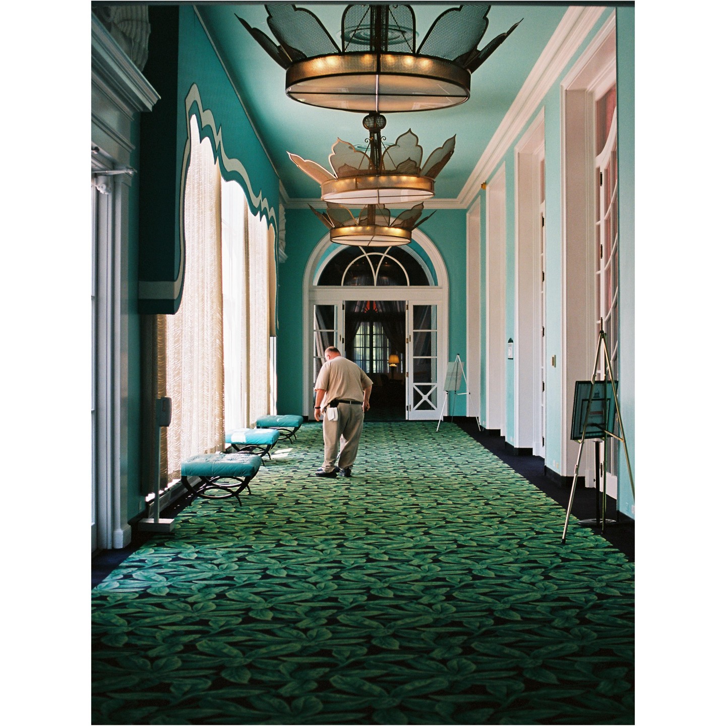 A man walks down a long hallway with a green leaf-patterned carpet and turquoise walls.