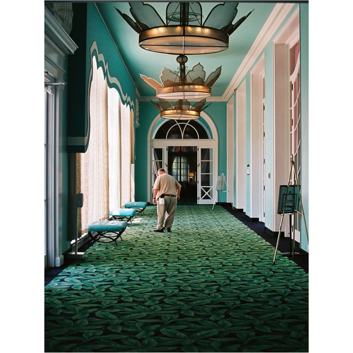 A man walks down a long hallway with a green leaf-patterned carpet and turquoise walls.