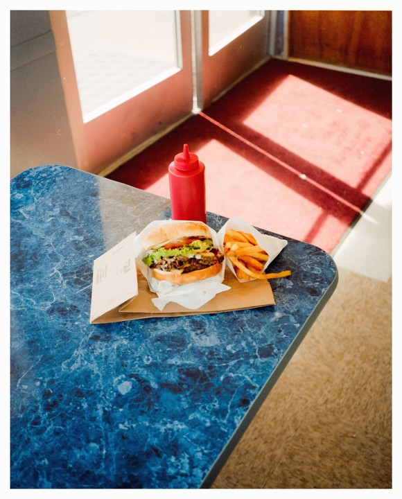 A burger and fries sit on a blue marbled table next to a red ketchup bottle, with sunlight streaming through a doorway in the background.