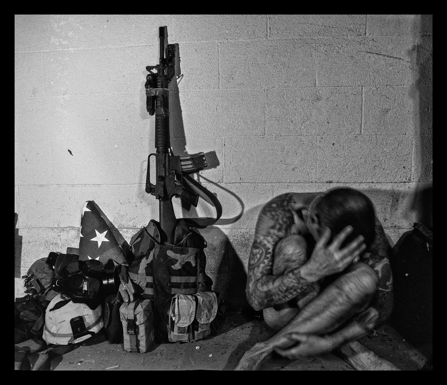 A man with tattoos is sitting on the ground next to a rifle and military gear.