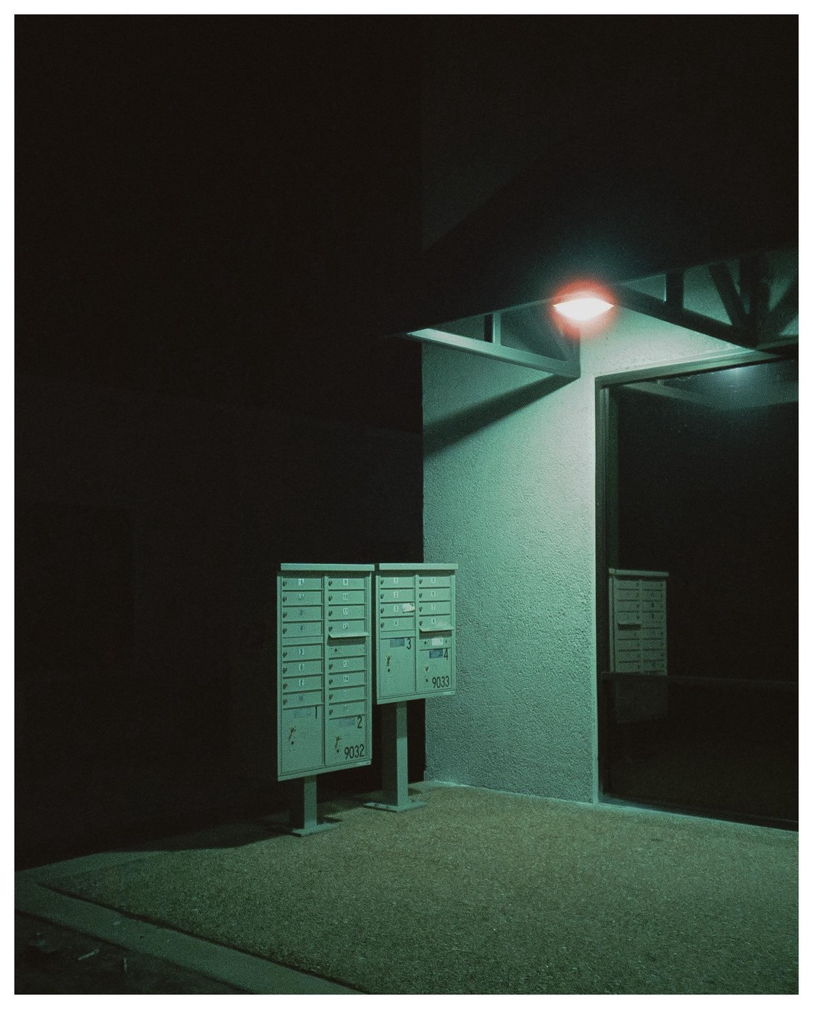 Two mailboxes stand on a textured ground next to a building with a glowing light at night.