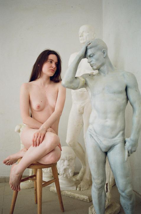 A nude woman sits on a wooden stool next to several statues of men.