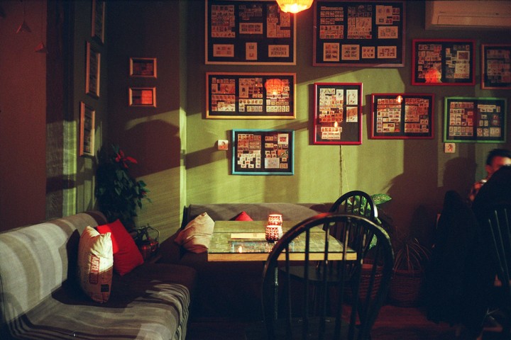 A cozy cafe interior features framed collections of stamps on a green wall, with a sofa, table, and chairs in the foreground.