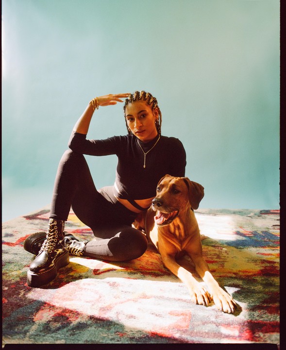 A woman with braided hair sits on a colorful rug next to a brown dog, both looking towards the viewer.