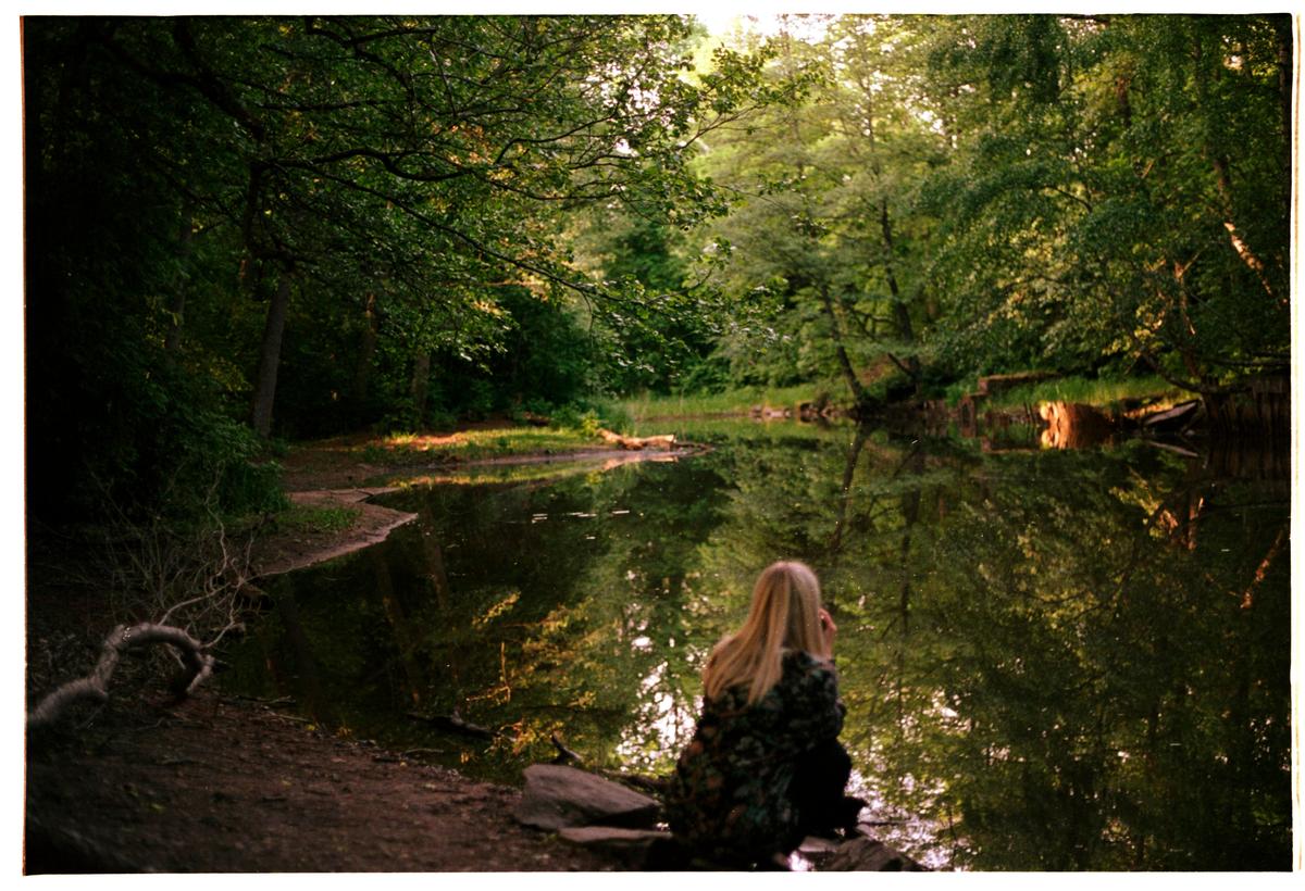 A woman sits by a still body of water reflecting the surrounding forest.