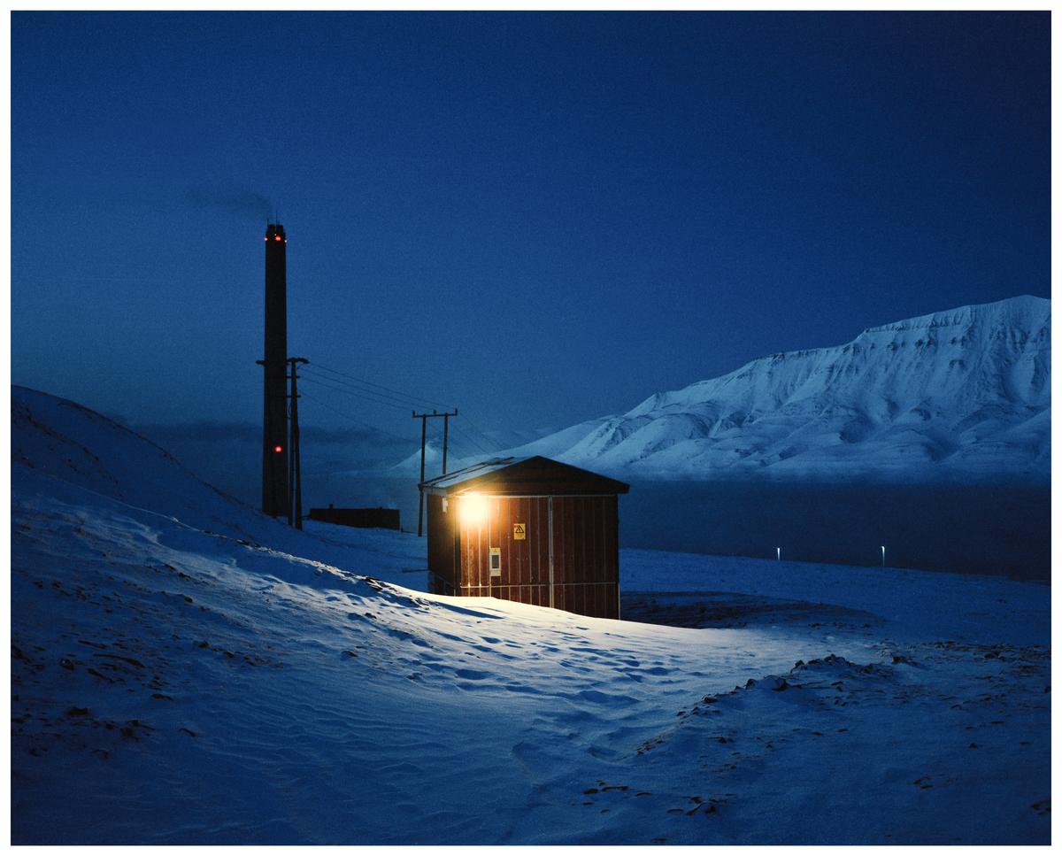 A small building with an illuminated window stands in a snow-covered landscape with a tall chimney and mountains in the background at night.