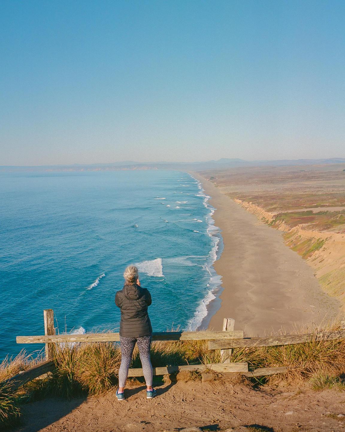 A woman stands on a cliff overlooking a long sandy beach and the ocean.