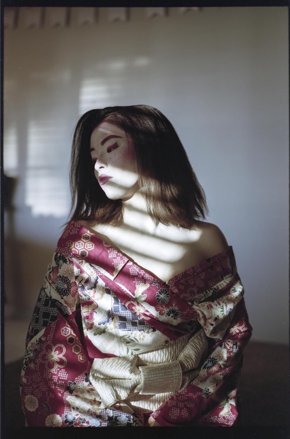 A woman with dark hair and dramatic makeup wears a colorful kimono with a white obi, illuminated by striped shadow.