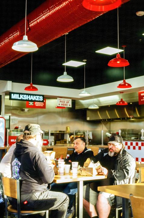 Four men are seated at a table in a restaurant, eating burgers and fries under red and white hanging lamps.