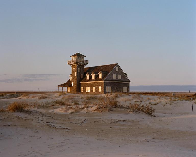 A historic lifesaving station with a tower stands on a sandy dune landscape with a bridge in the background.