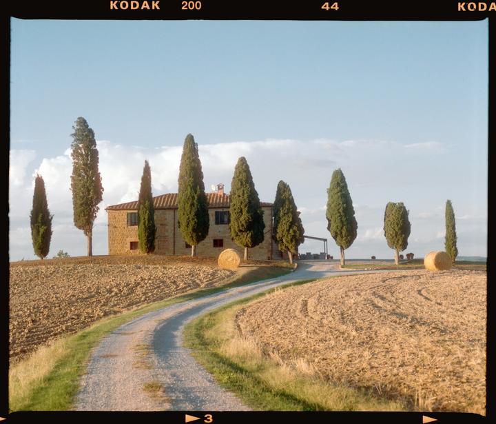 A winding dirt road leads to a stone house surrounded by cypress trees and hay bales in a field under a blue sky.
