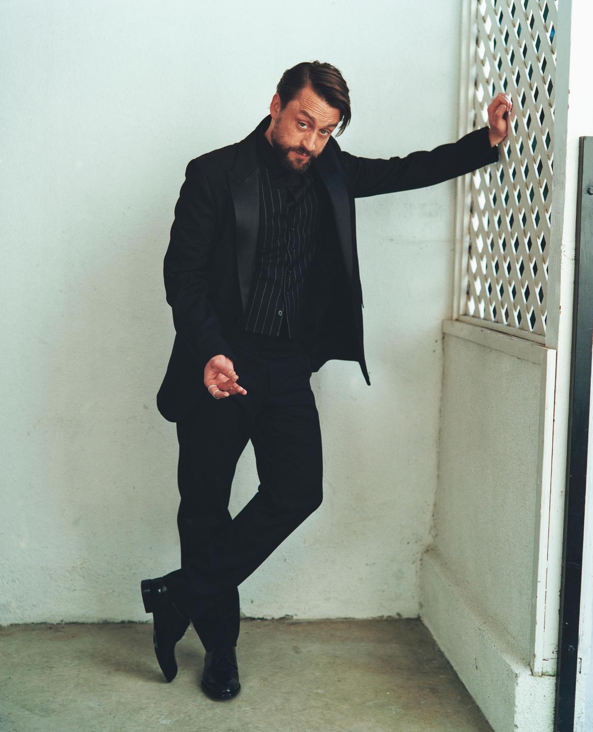 A man in a black suit and striped shirt leans against a white wall with a decorative screen.
