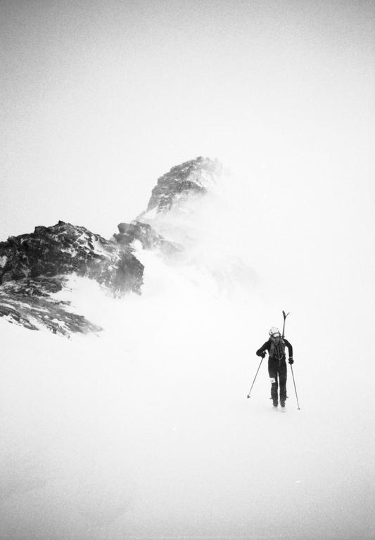A skier ascends a snowy mountain with skis strapped to their backpack, surrounded by a hazy, mountainous landscape.
