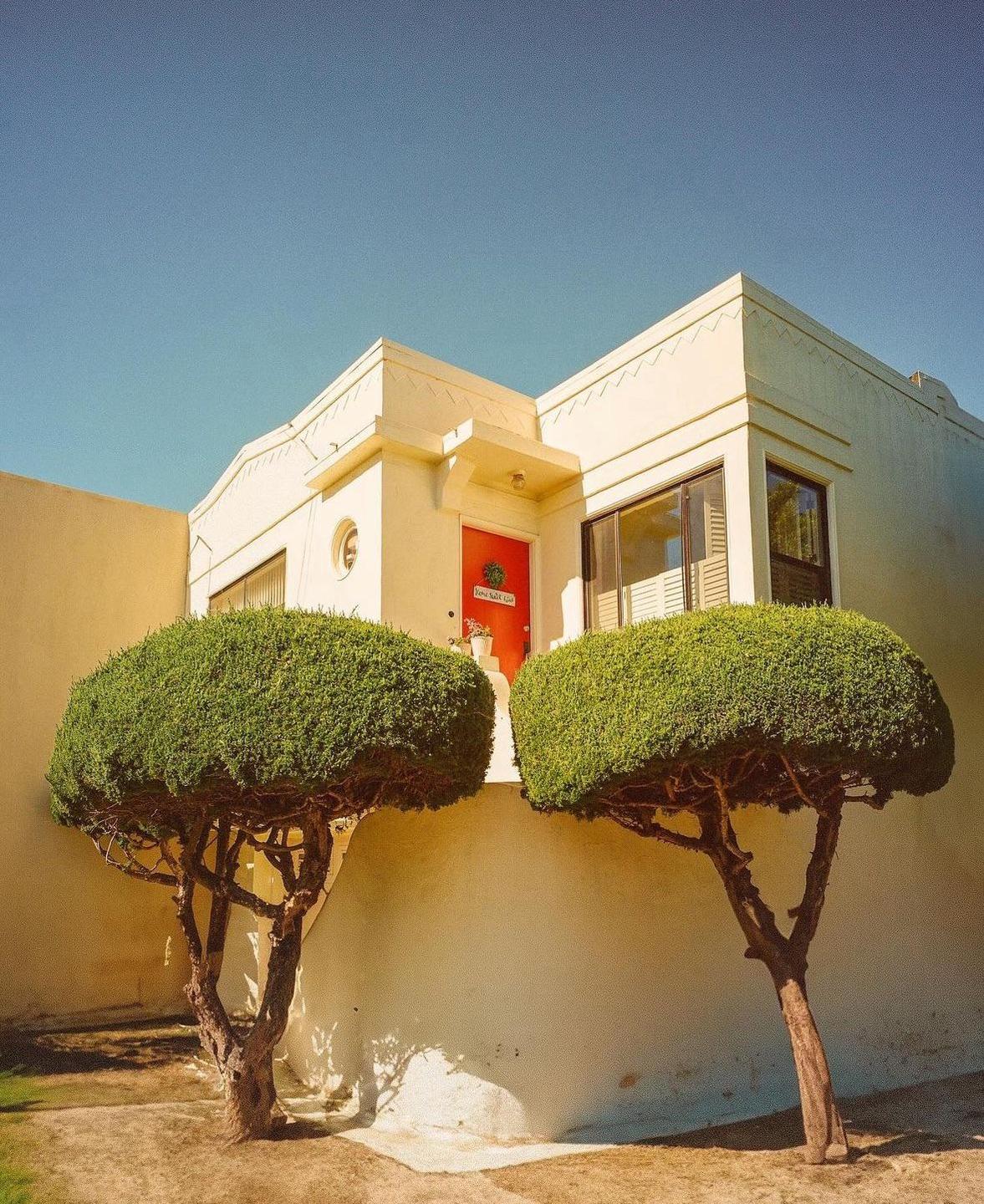 Two manicured trees stand in front of a cream-colored building with a red door.